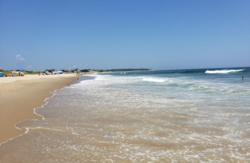 A sunny beach scene with gentle waves, soft sand, and a few people enjoying the water and shoreline.