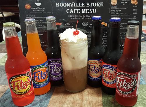 A glass of root beer float topped with whipped cream and a cherry, surrounded by colorful Fitz's soda bottles.