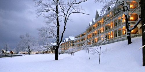 A snowy landscape featuring a large, illuminated building with balconies, surrounded by bare trees and a winter sky.