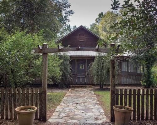 A rustic house surrounded by greenery, with a stone pathway leading to a wooden gate and potted plants on either side.
