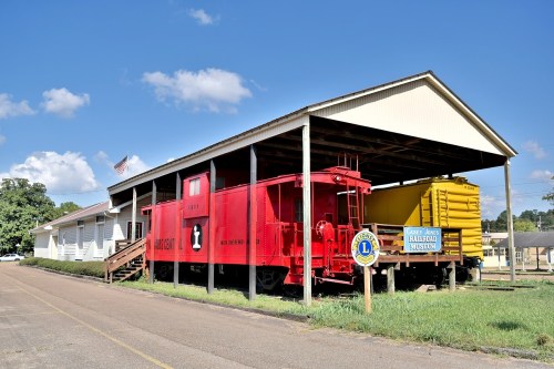 A red caboose and yellow train car under a shelter, next to a railroad museum building on a sunny day.