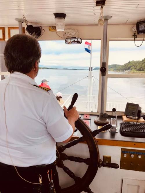 A captain steers a boat from the helm, with a flag visible outside and a scenic view ahead.
