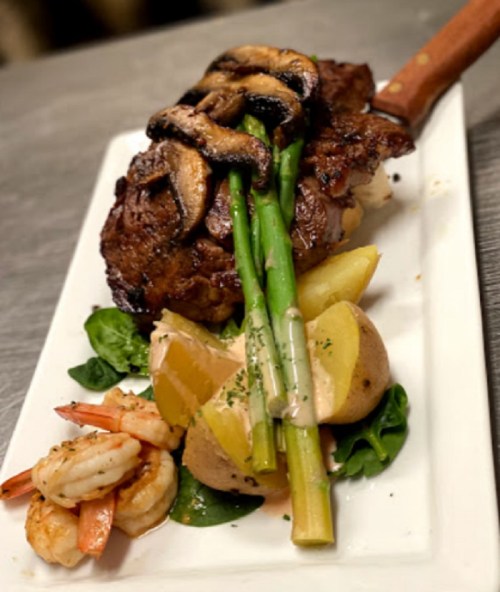 A gourmet dish featuring grilled steak topped with mushrooms, asparagus, potatoes, shrimp, and a creamy sauce on a white plate.
