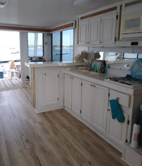 Bright interior of a boat featuring a white kitchen with cabinets, a gas stove, and a view of the water outside.