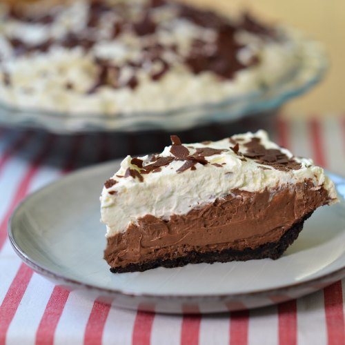 A slice of chocolate cream pie with whipped cream and chocolate shavings on a plate, with the whole pie in the background.