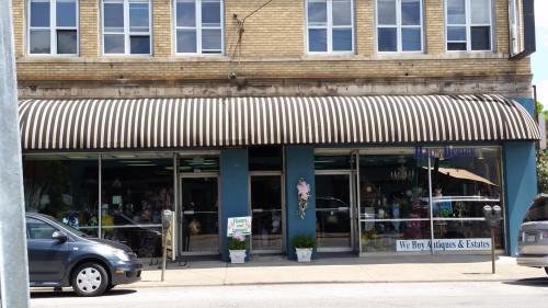 A storefront with a striped awning, displaying "We Buy Antiques & Estates" and various decorative items in the windows.