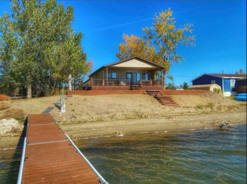 A lakeside cabin with a wooden dock, surrounded by trees and autumn foliage, on a sunny day.