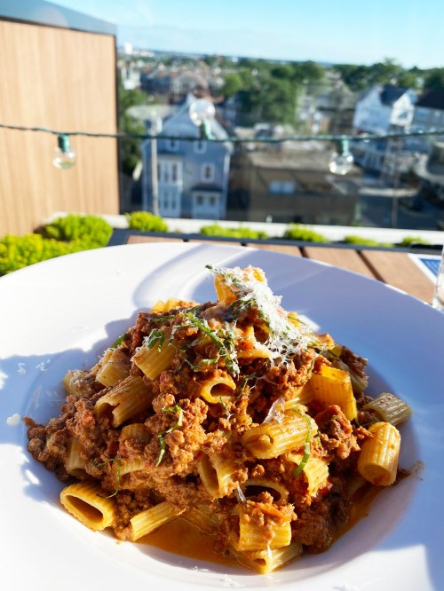 A plate of rigatoni pasta topped with meat sauce and grated cheese, with a scenic view in the background.