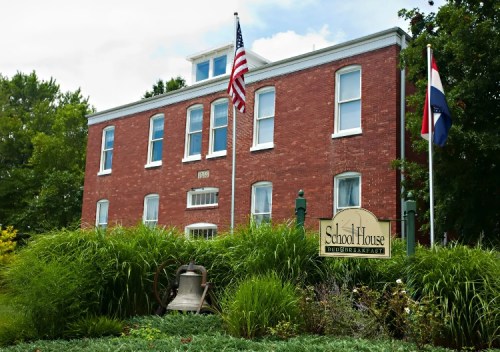Historic brick building with an American flag, a sign reading "School House," and lush greenery in front.