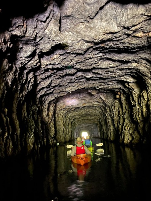 Two kayakers navigate through a dark, rocky cave with a bright light at the end, reflecting on the water.