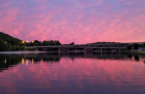 A serene river scene at sunset, reflecting pink and purple clouds with a bridge in the background.