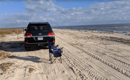 A parked SUV beside a beach with a single folding chair on the sandy shore and calm ocean waves in the background.