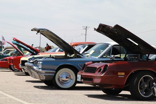 A row of classic cars with hoods open, showcasing their engines at a car show under a clear sky.