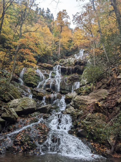 A serene waterfall cascades down rocky terrain, surrounded by vibrant autumn foliage and a tranquil pool below.
