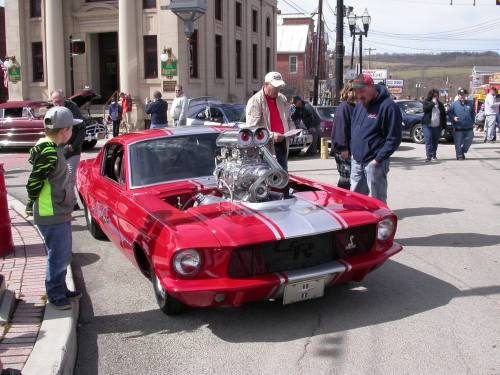 A red vintage car with a large engine is parked on a street, surrounded by onlookers and classic cars.