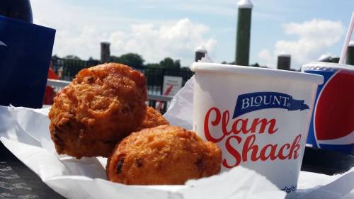 Two golden-brown clam fritters on a paper tray next to a cup labeled "Clam Shack" against a sunny outdoor backdrop.