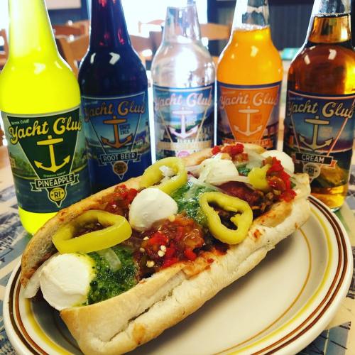A colorful hot dog topped with salsa, mozzarella, and peppers, alongside various Yacht Club soda bottles.