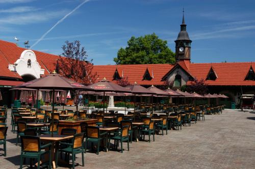 A sunny outdoor dining area with empty tables and umbrellas, set against a building with a clock tower.