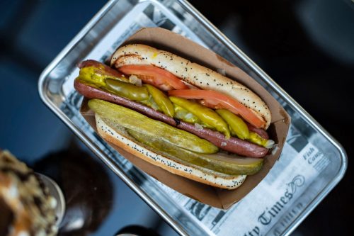 A hot dog in a poppy seed bun topped with pickles, tomatoes, and mustard, served on a tray.