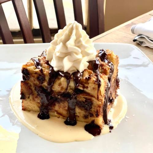 A slice of bread pudding topped with whipped cream and drizzled with chocolate sauce on a plate.