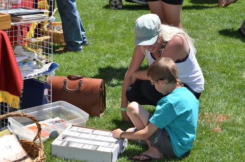 A woman and a child examine items in a box at an outdoor market on a sunny day.