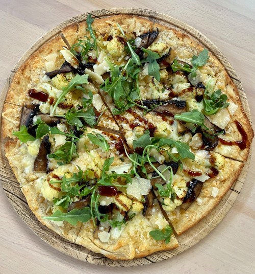 A gourmet pizza topped with mushrooms, cauliflower, greens, cheese, and drizzled with sauce on a wooden board.