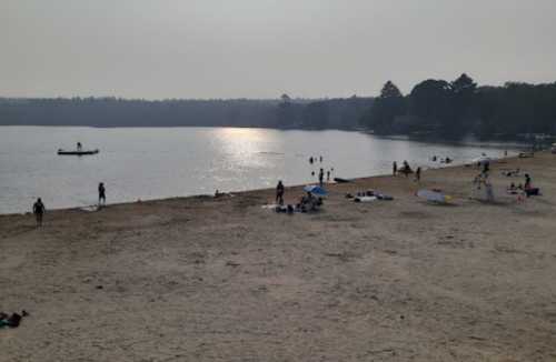 A sandy beach with people swimming, sunbathing, and a boat on the water under a hazy sky.