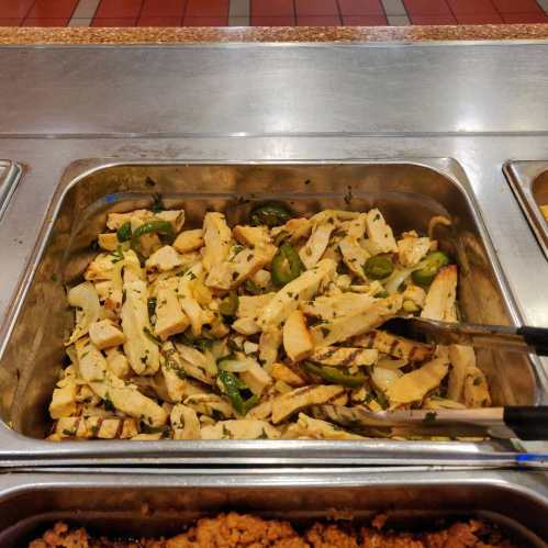 Grilled chicken strips with green peppers and herbs in a metal serving tray. Tongs are placed inside for serving.