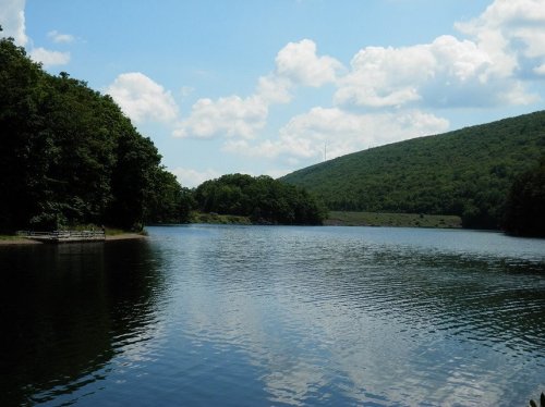 A serene lake surrounded by lush green trees and rolling hills under a partly cloudy sky.