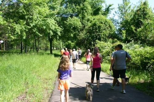 A group of people walking on a path through a green park, with a girl and a dog in the foreground.