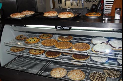 A display case filled with various pies, including fruit, nut, and cream varieties, in a bakery setting.