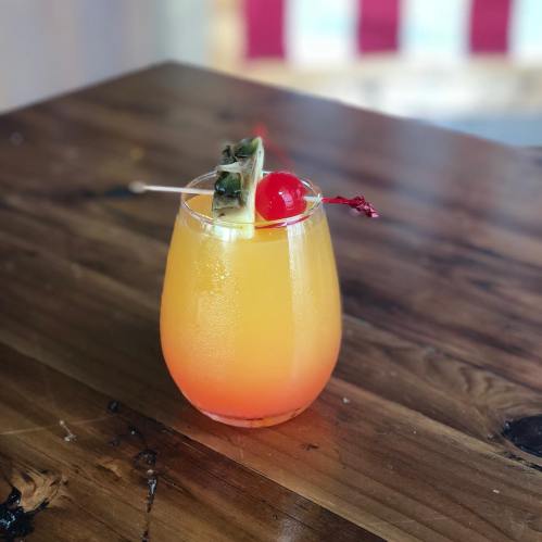 A colorful cocktail with orange and yellow layers, garnished with a cherry and pineapple, on a wooden table.