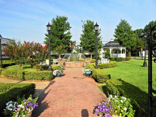 A landscaped garden with a fountain, gazebo, and flower beds, surrounded by trees and lampposts under a clear blue sky.