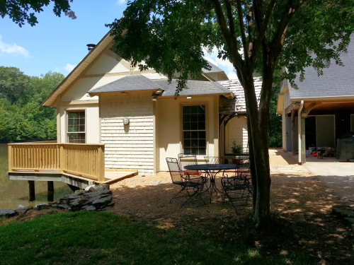 A cozy house with a wooden deck by a pond, surrounded by trees and a patio set on a gravel area.