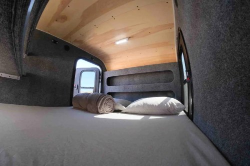 Cozy interior of a camper van with a bed, pillows, and a blanket, featuring wooden walls and a window.