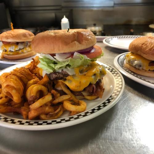 A juicy cheeseburger with lettuce, tomato, and onion, served with curly fries on a plate. Other burgers in the background.