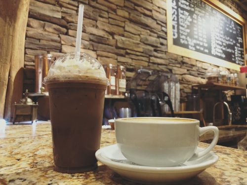A frosty iced drink and a steaming cup of coffee sit on a granite countertop in a cozy café setting.