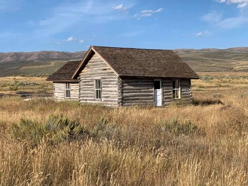 A rustic log cabin surrounded by tall grass and rolling hills under a blue sky.