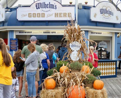 A food stand with a sign reading "Wilhelm's" and a fall-themed display featuring pumpkins and hay. People are in line.