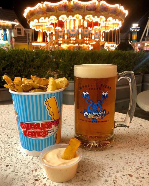 A cup of fries with dipping sauce and a beer mug, with a carousel lit up in the background at a festive fair.