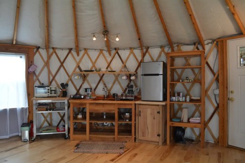 Cozy kitchen in a yurt, featuring wooden shelves, a fridge, and various appliances against a circular, wooden wall.
