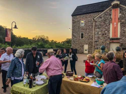 A lively outdoor gathering at the Hunterdon Art Museum, featuring food, drinks, and guests enjoying the sunset.