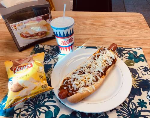 A plate with a chili cheese hot dog, a bag of Lay's chips, and a drink on a table with a floral placemat.