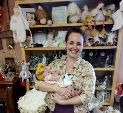 A smiling woman holds a newborn baby in a cozy shop filled with stuffed animals and toys.