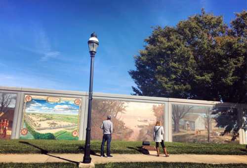 Two people stand in front of a large mural depicting scenic landscapes, with trees and a lamp post nearby.