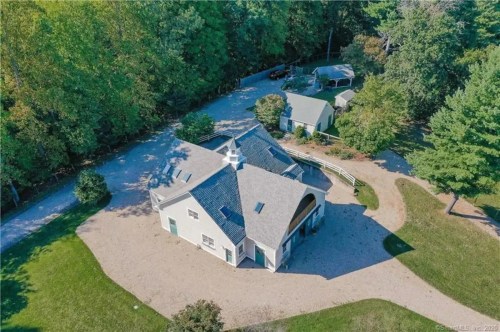 Aerial view of a circular house surrounded by trees and a gravel driveway, with additional buildings nearby.