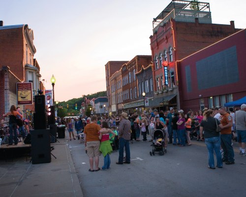 A lively street festival with a crowd enjoying music, food, and activities as the sun sets over historic buildings.