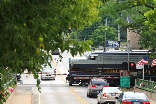 A train passes through a level crossing, with cars stopped and trees framing the scene. Nearby signs are visible.