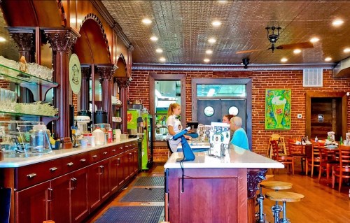 A cozy café interior with a wooden counter, brick walls, and two customers chatting near the register.