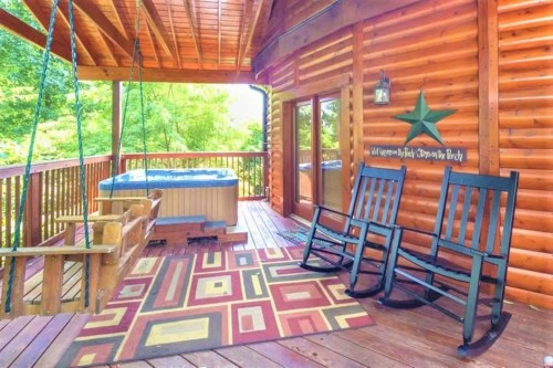 Cozy wooden deck with rocking chairs, a swing, and a hot tub, surrounded by greenery. Colorful rug adds warmth.